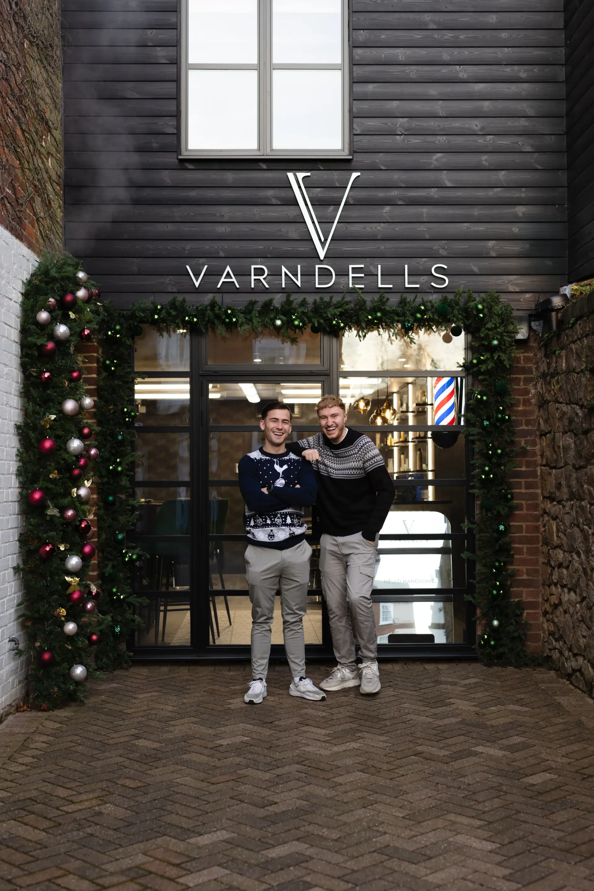 Front of Varndells barbershop with a dark wooden facade, festive green garlands, and colorful ornaments. Two men in Christmas sweaters stand smiling in front of the entrance, capturing a cheerful holiday atmosphere.