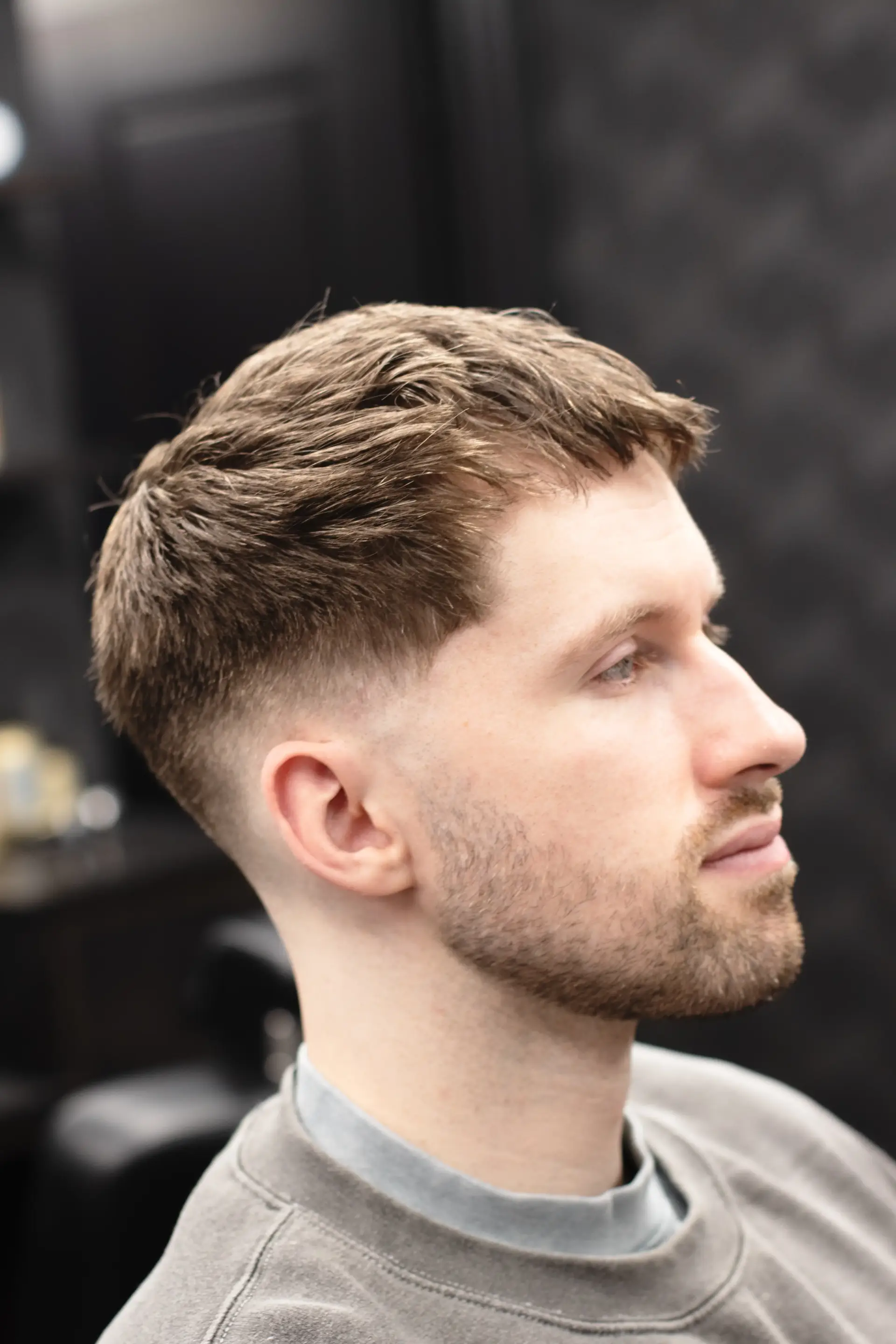 Angled front view of a man sporting short, textured brown hair and a neatly shaped beard, emphasizing a low fade and casual grey sweatshirt in a contemporary barbershop setting.
