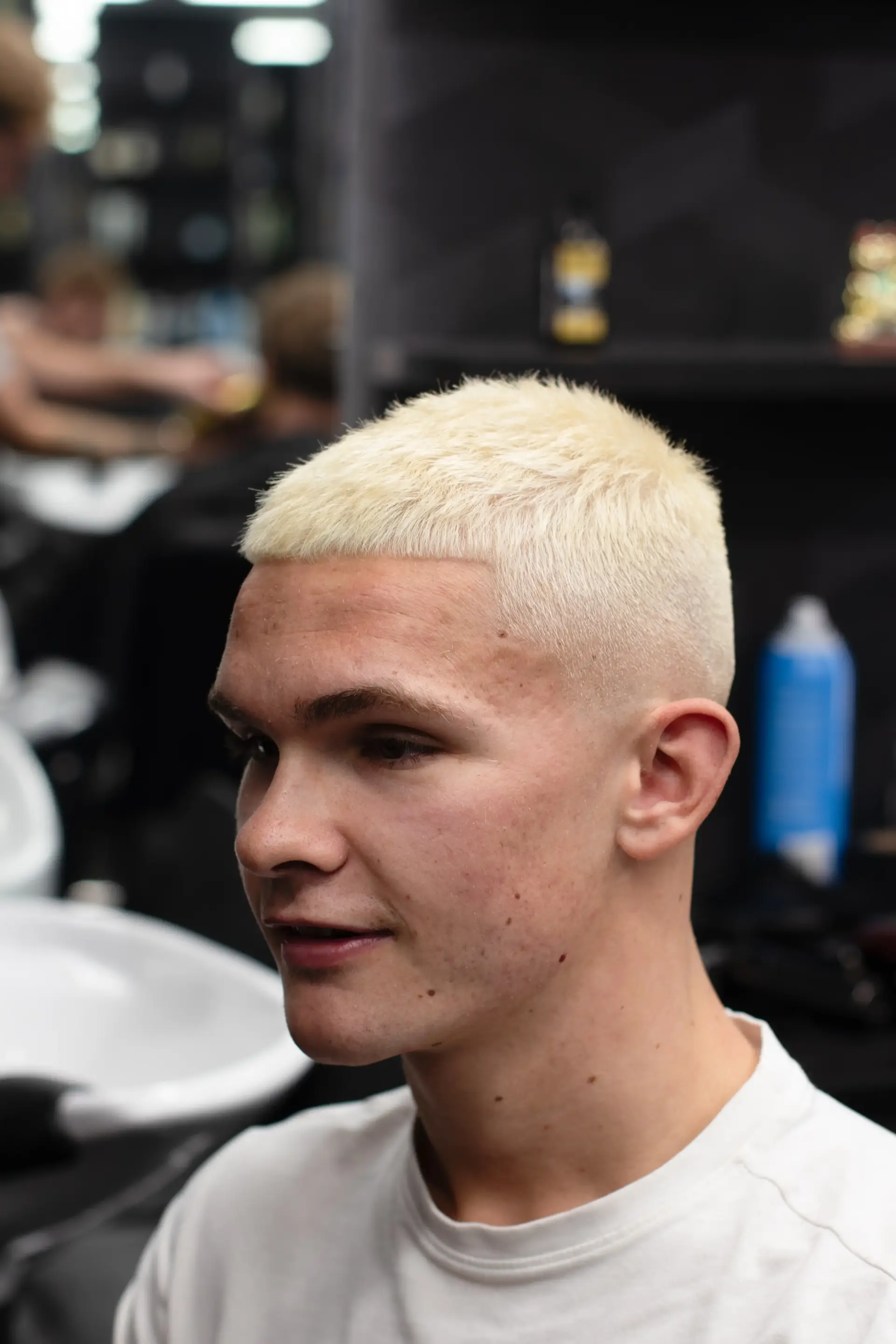 Close-up of a young man with a crisp platinum-blonde buzz cut, wearing a white T-shirt and sitting in a modern barbershop