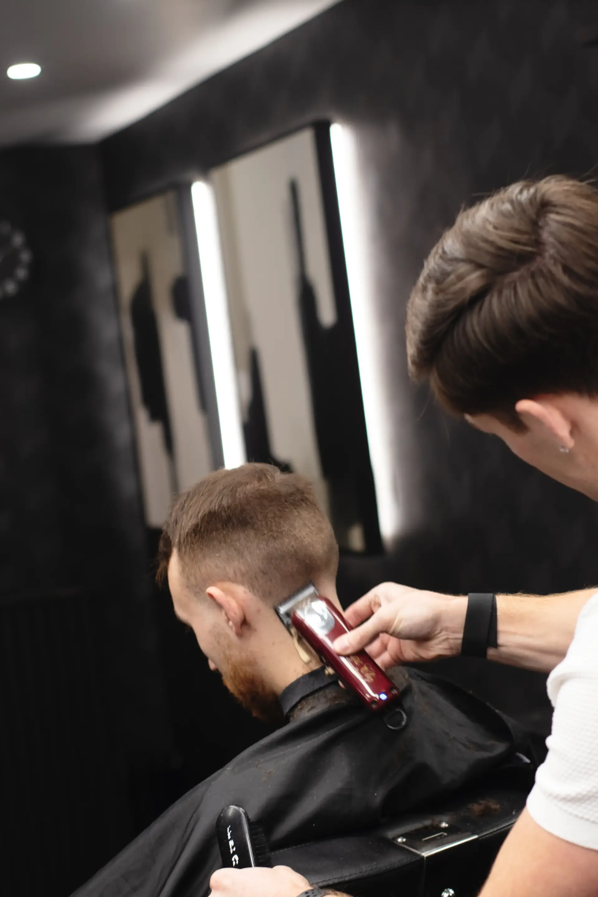 Rear view of a man in a black cape getting a neatly tapered fade from a barber using a red hair clipper, set against a modern barbershop interior.