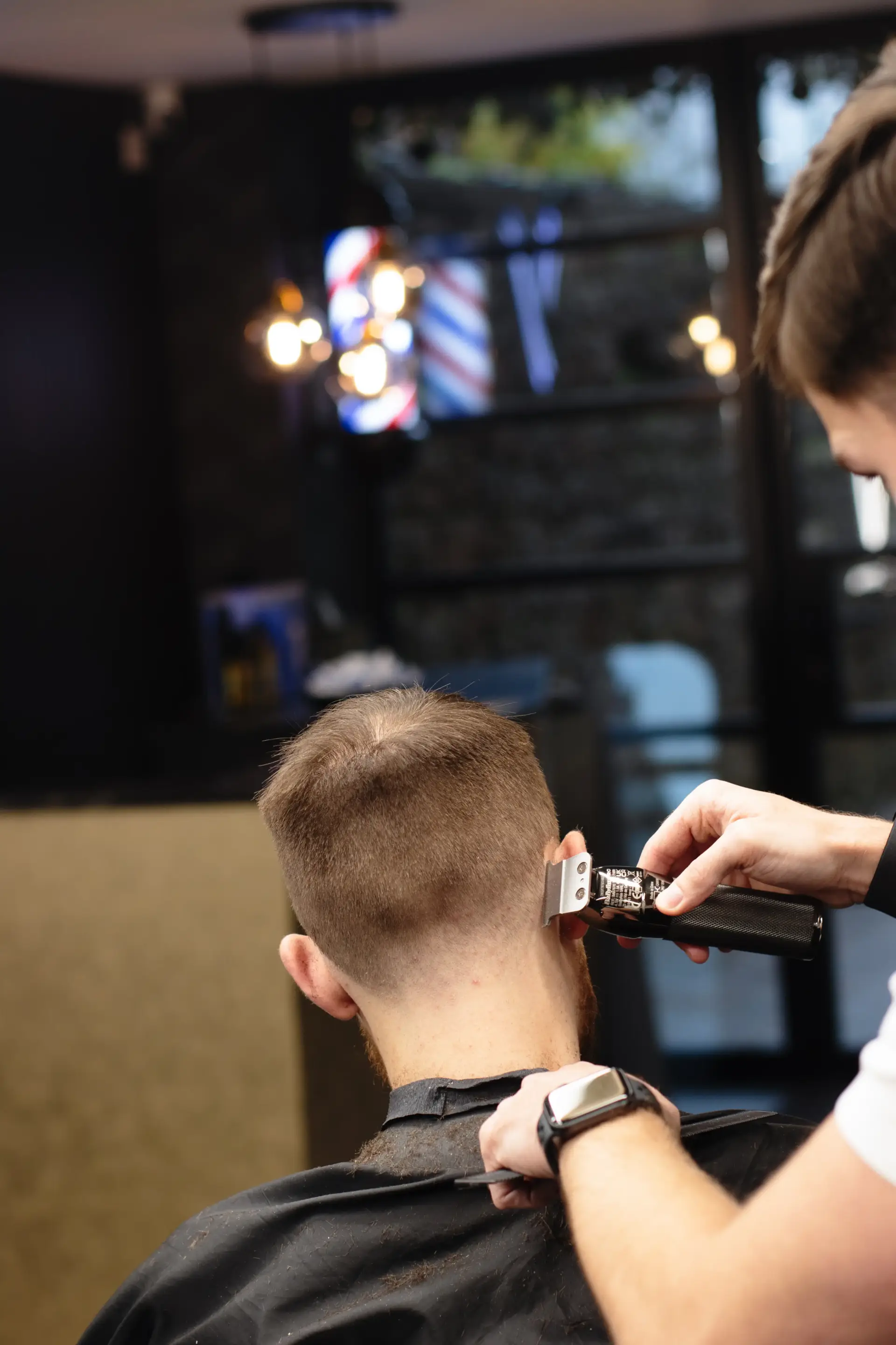 Barber refining the back of a man’s fade haircut using a silver trimmer, with the client in a black cape and a stylish barbershop in the background.