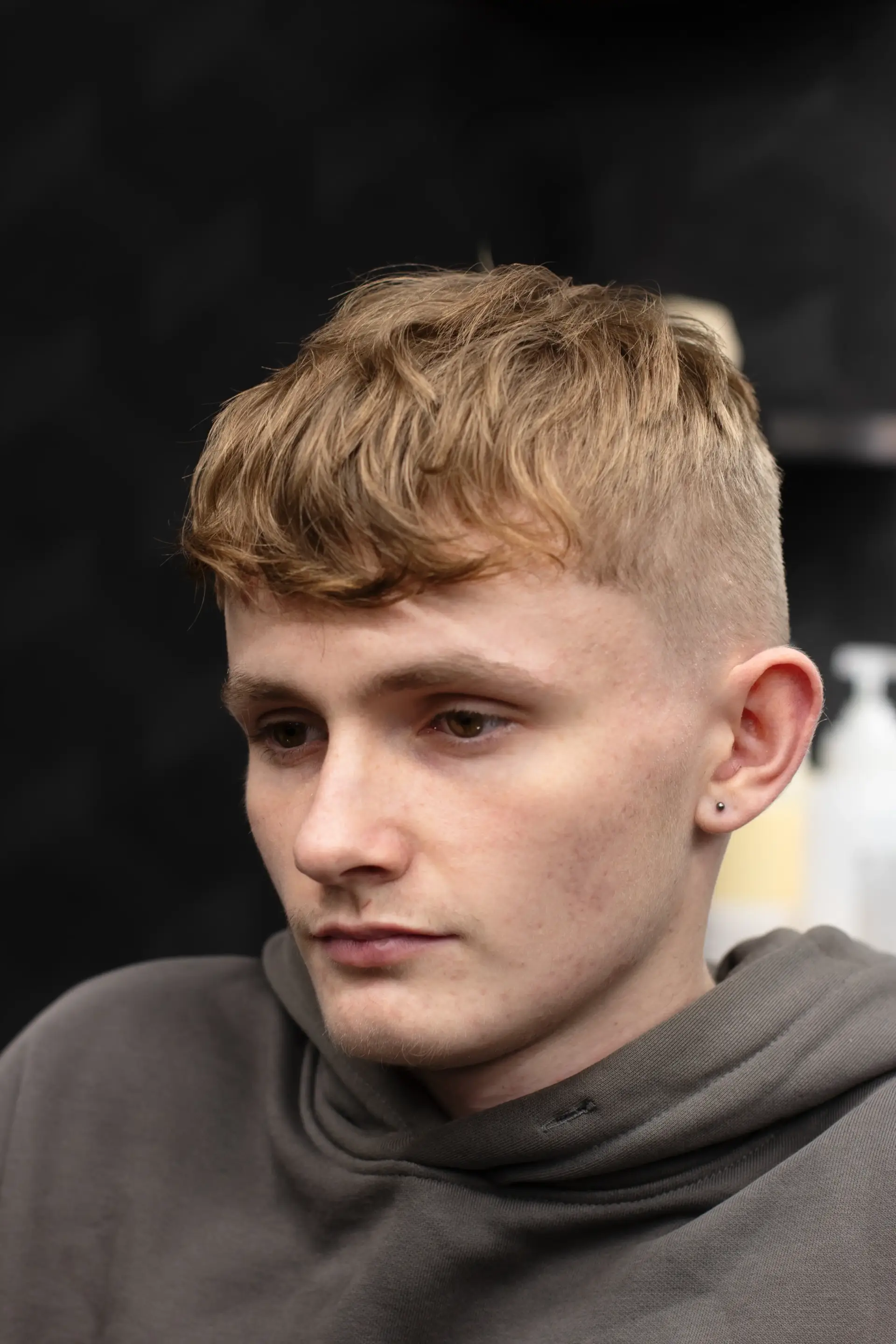 Front-angled close-up of a young man in a grey hoodie, highlighting his short, layered blonde crop haircut and neatly faded sides in a contemporary salon setting.