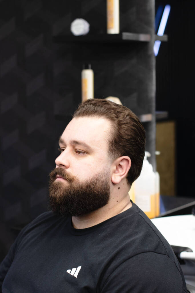 Profile view of a man with slicked-back medium-length hair and a sharply trimmed beard, seated in a stylish barbershop.