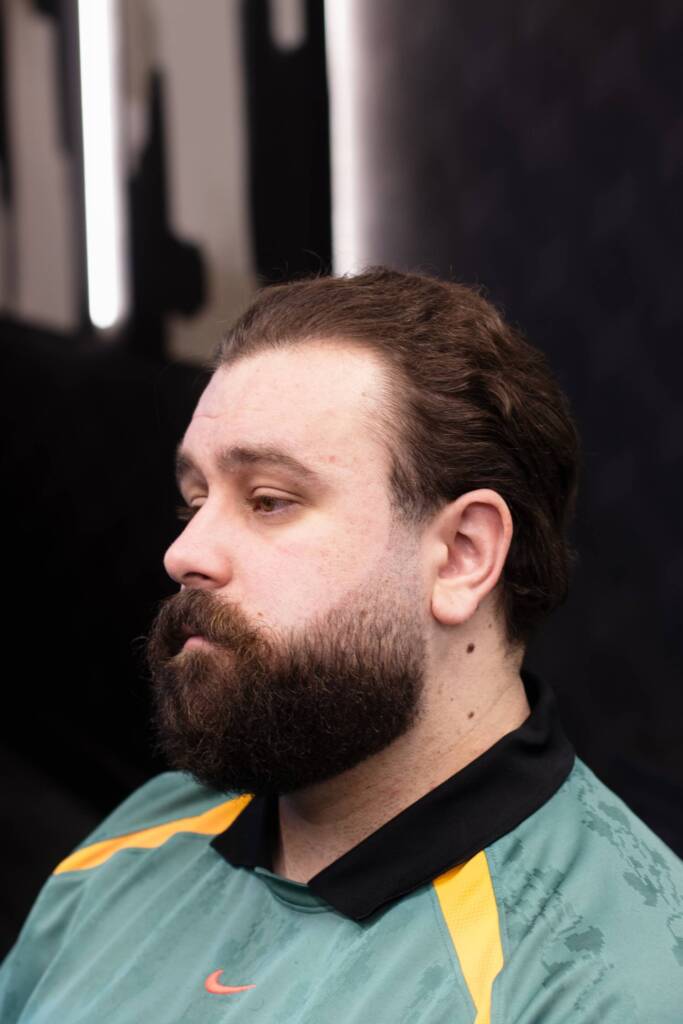 Side profile of a man growing out mid-length hair with a full beard, wearing a green and orange sports top, taken inside a luxury barbershop.