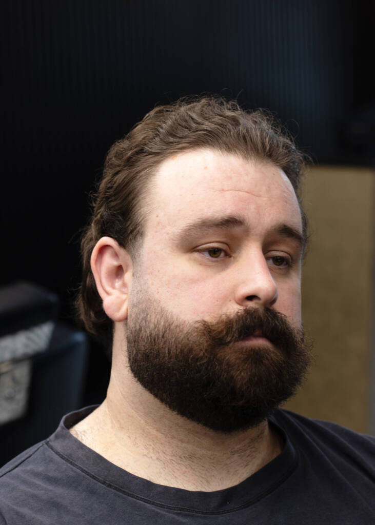 Man with a natural, brushed-back medium-length hairstyle and thick beard with curled moustache, seated inside a barbershop.